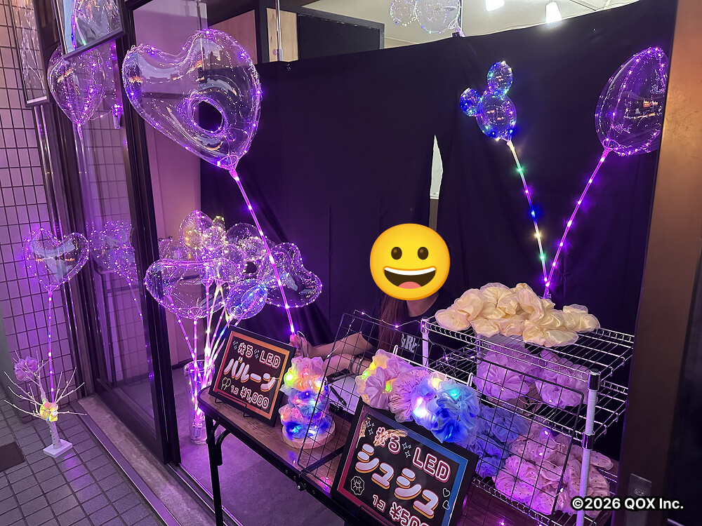 光るバルーンとシュシュを飾った店頭の机に女性スタッフが座って販売する様子